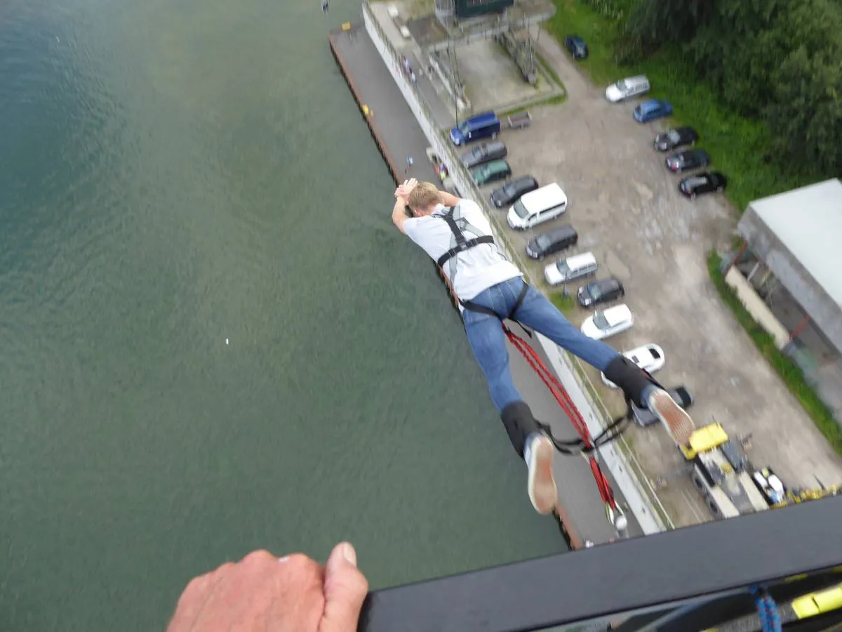 Bungeespringer in weißem T-Shirt und Jeans springt von oben, mit Blick auf das Wasser.